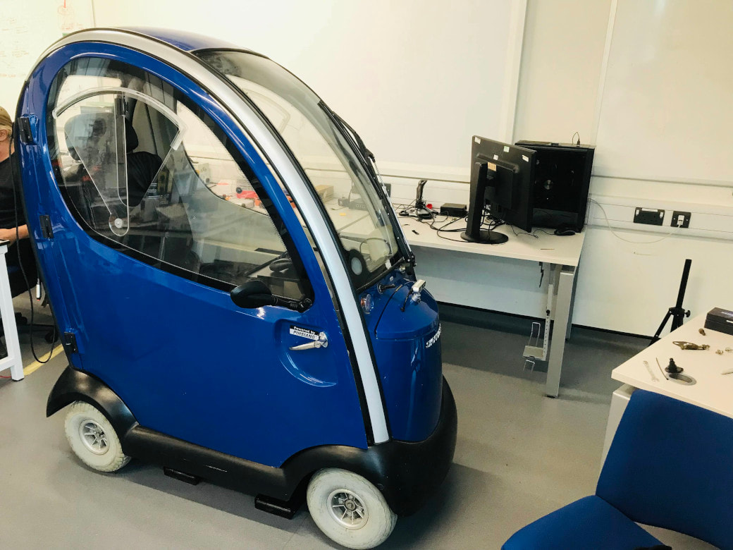 The OpenPodCar_V2 vehicle, a converted blue hard-canopy mobility scooter, in the lab